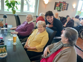 Eine Gruppe älterer Menschen sitzt an einem Tisch in einem Speisesaal. Eine Betreuerin steht lächelnd hinter einer Frau in gelber Bluse. Auf dem Tisch stehen Getränke und Snacks. Im Hintergrund sind weitere Bewohner zu sehen.