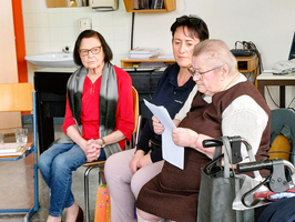 Drei Frauen sitzen in einem Raum. Die linke Frau trägt eine rote Bluse und eine dunkle Jacke, die mittlere hat ein schwarzes Oberteil an, und die rechts sitzt mit einem Rollator. Die rechte Frau liest einen Text. Im Hintergrund sind Möbel und ein Computer sichtbar.