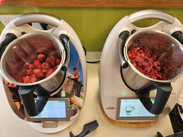 Zwei Küchengeräte stehen auf einer Arbeitsplatte. Das linke Gerät enthält frische Erdbeeren, das rechte gefrorene Himbeeren. Beide Geräte haben digitale Displays und sind in moderner, futuristischer Optik gestaltet.