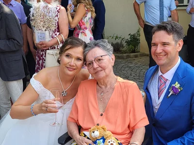 Brautpaar posiert mit einer Frau im Rollstuhl, umgeben von Hochzeitsgästen.