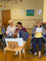 Eine Gruppe von älteren Frauen sitzt in einem Wohnraum. Eine Frau spielt ein Akkordeon, während die anderen Frauen Liedertexte in den Händen halten und singen. Im Hintergrund sind dekorative Bilder und Blumenarrangements zu sehen.