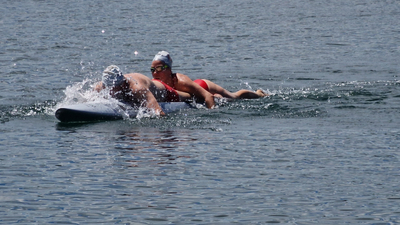 Zwei Schwimmer paddeln auf einem Board durch ruhiges Wasser.