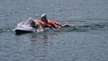 Zwei Schwimmer paddeln auf einem Board durch ruhiges Wasser.