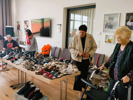 Eine Gruppe älterer Menschen betrachtet verschiedene Schuhe, die auf einem Tisch ausgestellt sind. Im Hintergrund sind einige Stühle und ein Fernseher zu sehen. Zwei Frauen stehen am Tisch, während ein Mann und eine weitere Frau im Hintergrund sitzen.