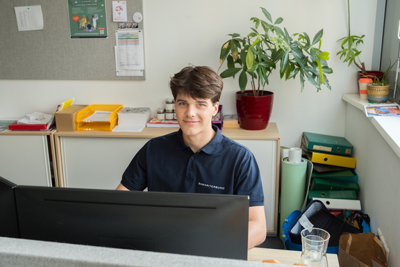 Zivildiener sitzt vor Computer, im Hintergrund Büro, Pflanzen