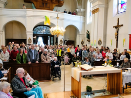 Eine große Gruppe von Menschen steht und sitzt in einer Kirche. Einige sind auf Rollstühlen, andere hören aufmerksam zu. Im Vordergrund sind ein Altar und Dekorationen sichtbar. Das Licht strömt durch die Fenster und schafft eine feierliche Atmosphäre.
