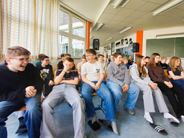 Eine Gruppe von Schülern sitzt in einem Klassenraum. Einige lachen, während andere amüsiert oder nachdenklich wirken. Im Hintergrund sind Fenster und eine Tafel sichtbar. Die Schüler tragen unterschiedliche Kleidungsstile und scheinen an einer Veranstaltung teilzunehmen.