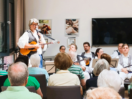 Eine Sängerin mit einer Gitarre steht im Vordergrund und spricht ins Mikrofon. Im Hintergrund sitzen mehrere Musiker, die Gitarre spielen, und ein Publikum, das aufmerksam zuhört. Der Raum hat große Fenster und einige Bilder an den Wänden.