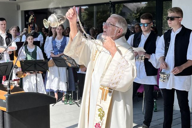 Ein Priester in einer weißen Robe schwenkt ein Weihrauchgefäß während einer Zeremonie auf der Terrasse vor einem Gebäude. Um ihn herum stehen mehrere Personen in traditioneller Kleidung. Manche halten Musikinstrumente für einen Auftritt in der Hand.