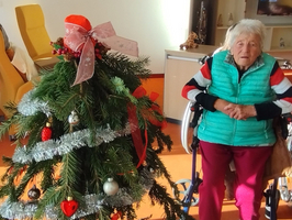 Eine ältere Frau sitzt neben einem geschmückten Weihnachtsbaum in einem Wohnraum.