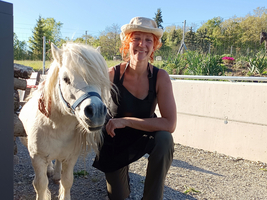 Eine Person mit orangefarbenen Haaren kniet neben einem kleinen weißen Pony. Die Person trägt ein schwarzes Oberteil und eine helle Hut. Im Hintergrund sind Pflanzen und Bäume zu sehen, sowie ein blauer Himmel.