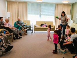 Eine Gruppe von Kindern steht in einem Raum und beobachtet eine Frau, die eine Aktivität leitet. Im Hintergrund sitzen ältere Menschen auf Stühlen. Die Atmosphäre wirkt freundlich und interaktiv, mit bunten Kleidungsstücken der Kinder und einem hellen, einladenden Raum.