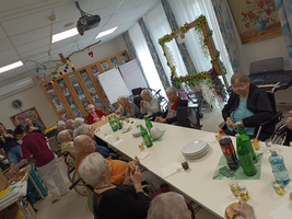 Eine Gruppe älterer Menschen sitzt an einem langen Tisch in einem Gemeinschaftsraum. Auf dem Tisch stehen Getränke und Speisen. Im Hintergrund sind einige Personen aktiv, während ein dekorierter Rahmen für Fotos aufgestellt ist. Das Ambiente wirkt freundlich und gesellig.