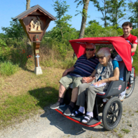 Ein älteres Paar sitzt zusammen in einem Rollstuhl, während ein junger Mann ihn schiebt.