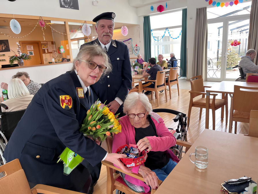 Frau in Uniform überreicht Blumen und Süßigkeiten an ältere Dame im Aufenthaltsraum.