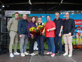 Eine Gruppe von sieben Menschen steht zusammen auf der Bühne vom Tag des Samariterbundes mit einem Blumenstrauß.