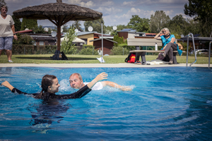 Ein Mann hilft einem Mädchen im Schwimmbecken, während eine Frau auf einer Liege im Hintergrund entspannt. Im Hintergrund sind weitere Personen und ein Garten zu sehen. Die Szene vermittelt eine entspannte, sommerliche Atmosphäre.