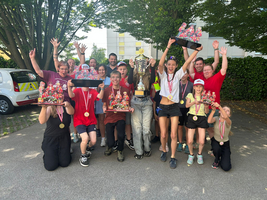 Eine Gruppe von Menschen, die in einem Outdoor-Bereich posieren, hebt ihre Hände in die Luft und lächelt. Einige halten Trophäen und Medaillen. Der Hintergrund zeigt Bäume und ein Auto. Die Atmosphäre ist feierlich und voller Freude.