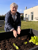 Eine ältere Dame steht an einem Hochbeet und lächelt. Sie hat die Hände über die Erde gelegt, in der kleine Pflanzen und Samen wachsen. Im Hintergrund ist ein Gebäude und eine grüne Wiese zu sehen.