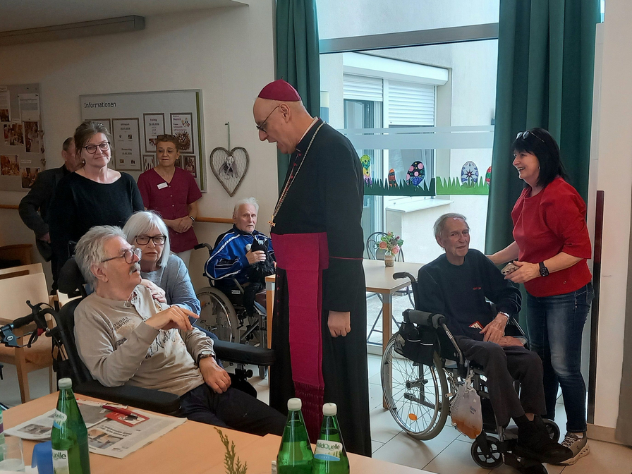 Eine Gruppe von Menschen in einem Gemeinschaftsraum. Der Bischof in religiöser Kleidung spricht mit einem älteren Mann im Rollstuhl. Weitere Personen, darunter Mitarbeiter und Senioren, stehen im Hintergrund und beobachten die Interaktion. Die Atmosphäre ist freundlich und familiär.