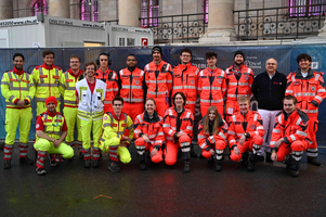 Eine Gruppe von Rettungskräften in orangefarbenen und gelben Uniformen steht gemeinsam draußen. Sie lächeln und posieren für das Foto. Im Hintergrund sind Baustellen und einige Fenster sichtbar. Die Atmosphäre wirkt freundlich und professionell.