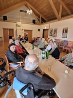 Eine Gruppe älterer Menschen sitzt an einem langen Tisch in einemSaals. Einige der Personen schauen zur Kamera, während sie miteinander sprechen. Auf dem Tisch stehen mehrere Flaschen Wasser und Gläser. Der Raum hat eine warme, einladende Atmosphäre.