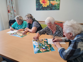 Vier ältere Frauen sitzen an einem Tisch und zeichnen oder schreiben. Sie konzentrieren sich auf bunte Bilder, die vor ihnen liegen. Im Hintergrund hängt ein großes Bild mit Blumenmotiven an der Wand.