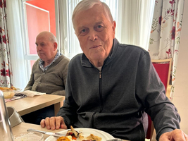 Ein älterer Mann sitzt an einem Tisch und genießt eine Mahlzeit, die auf einem Teller vor ihm serviert ist. Er trägt einen dunkelgrauen Pullover und schaut direkt in die Kamera. Im Hintergrund ist ein weiterer Mann und eine gemütliche Essensatmosphäre zu sehen.