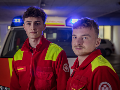 Zwei junge Sanitäter in roten Uniformen mit reflektierenden Elementen stehen vor einem Krankenwagen mit eingeschaltetem Blaulicht. Sie blicken ernst in die Kamera, während im Hintergrund die Umrisse eines Parkplatzes sichtbar sind.
