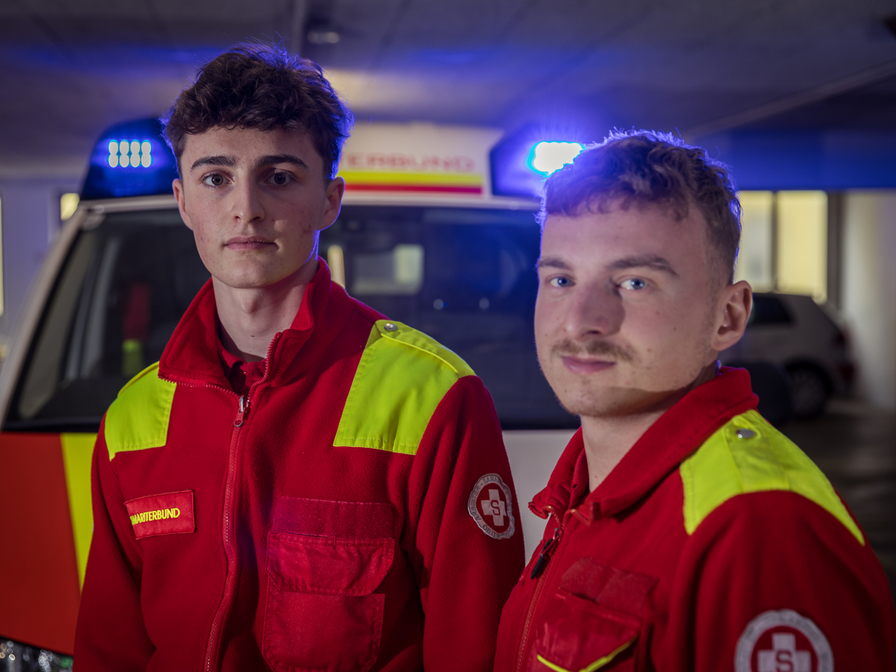Zwei junge Sanitäter in roten Uniformen mit reflektierenden Elementen stehen vor einem Krankenwagen mit eingeschaltetem Blaulicht. Sie blicken ernst in die Kamera, während im Hintergrund die Umrisse eines Parkplatzes sichtbar sind.