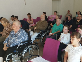 Eine Gruppe von Senioren und Kindern sitzt in einem Raum und hört aufmerksam zu.
