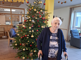 Ältere Frau mit Rollator vor einem festlich geschmückten Weihnachtsbaum.