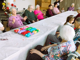 Eine Gruppe von älteren Menschen sitzt um einen langen Tisch und spielt mit bunten Plastikbällen. Einige Personen lächeln und interagieren, während eine Person im Hintergrund Winken zeigt. Der Tisch ist mit einer weißen Tischdecke bedeckt.
