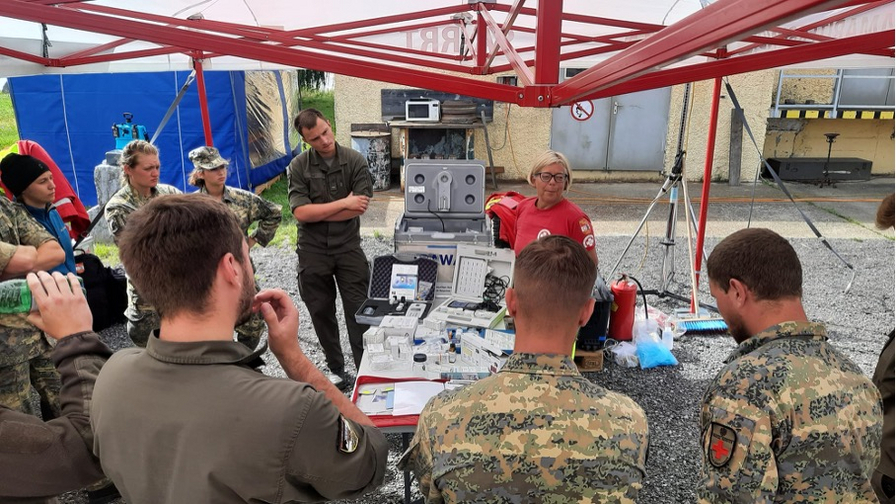 Eine Gruppe von Personen hört einer Ausbilderin zu, die medizinisches Equipment erklärt.
