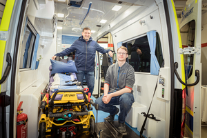 Zwei Männer stehen in einem Krankenwagen, einer sitzt auf der Trage.