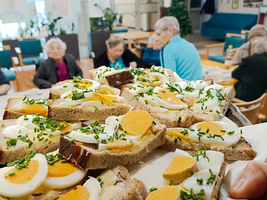 Ein Tablett mit belegten Broten, die mit Ei, Mayonnaise und Schnittlauch garniert sind. Im Hintergrund sind ältere Menschen an Tischen zu sehen, die miteinander sprechen. Die Umgebung wirkt freundlich und einladend.