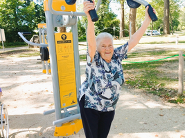 Ältere Frau trainiert an einem Fitnessgerät im Freien und lächelt.