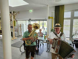 Zwei Musiker stehen im Vordergrund und spielen ein Tuba und ein Akkordeon. Im Hintergrund sitzt eine Person an einem Tisch in einem hellen Raum mit großen Fenstern und grünen Vorhängen. Die Atmosphäre ist festlich und einladend.