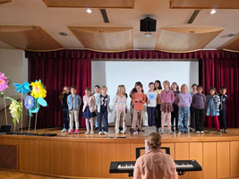 Kinder stehen auf einer Bühne und singen, während jemand Klavier spielt.