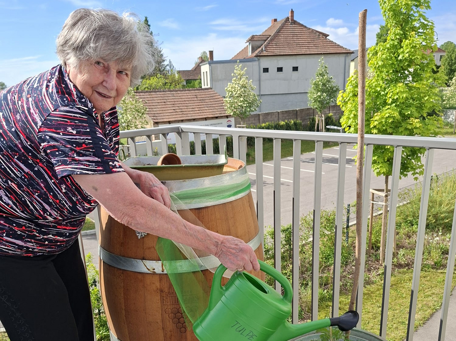 Eine ältere Frau mit grauen Haaren steht auf einem Balkon und gießt mit einer grünen Gießkanne Pflanzen in einem Blumentopf. Neben ihr steht ein Holzfass. Im Hintergrund sind Häuser und Bäume zu sehen.