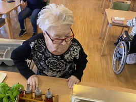 Ältere Frau bereitet Zutaten in einer Küche vor, während andere im Hintergrund sitzen.
