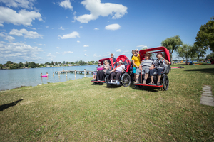 Ältere Menschen auf einer Wiese am Wasser in roten Rikschas sitzend.