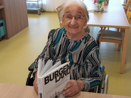 Eine lächelnde ältere Dame sitzt im Rollstuhl und hält ein Buch mit dem Titel „BURGEN“.