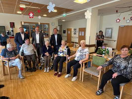 Eine Gruppe von Senioren sitzt in einem Gemeinschaftsraum. Einige stehen hinter ihnen. Die Einrichtung ist freundlich dekoriert, mit Blumen und einem warmen Holzfußboden. Die Personen scheinen an einer sozialen Veranstaltung teilzunehmen.