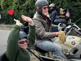 Zwei Personen auf einem Motorrad, eine Frau in einem Seitenwagen mit einer Brille und einer Kopfbedeckung, während ein Mann mit einer Sonnenbrille und einem Hut fährt. Beide sind sichtbar begeistert und lächeln, umgeben von einer grünen Landschaft.
