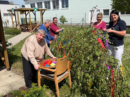 Eine Gruppe von fünf Personen arbeitet im Freien in einem Garten. Sie dekorieren ein mit bunten Stoffstücken geschmücktes, mit Zweigen versehenes Möbelstück. Der Himmel ist klar, und im Hintergrund sind Bäume sowie ein Gebäude zu sehen.