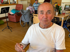 Ein älterer Mann sitzt an einem Tisch und isst. Er trägt ein weißes T-Shirt und hält eine Gabel in der Hand. Auf dem Teller vor ihm sind Reste von Essen. Neben ihm steht eine Tasse, und im Hintergrund sind weitere Personen und Möbel zu sehen.