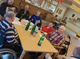 Eine Gruppe älterer Menschen sitzt an einem Tisch in einem Gemeinschaftsraum. Sie lächeln und unterhalten sich, während einige Wasserflaschen auf dem Tisch stehen. Im Hintergrund ist eine festliche Dekoration zu sehen.
