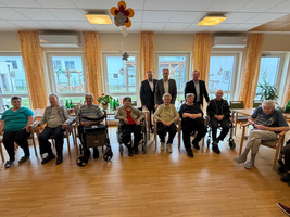 Gruppenfoto in einem Wohnheim mit mehreren älteren Menschen, die in Rollstühlen oder auf Stühlen sitzen. Im Hintergrund stehen zwei Männer in Anzügen. Der Raum ist hell und freundlich mit großen Fenstern und Dekorationen.