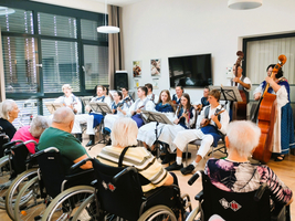 Eine Gruppe von Musikern spielt auf Instrumenten für ältere Menschen in Rollstühlen. Die Musiker tragen traditionelle Kleidung und sitzen in einem hellen Raum mit einem Fernseher im Hintergrund. Die älteren Menschen hören aufmerksam zu.